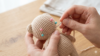 Mãos de uma artesã utilizando uma agulha de tapeceiro prateada para costurar cuidadosamente a cabeça de um pequeno urso amigurumi bege ao seu corpo, com alfinetes coloridos marcando a posição.