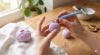 Mãos delicadas segurando uma agulha de crochê ergonômica com cabo de silicone e um novelo de fio de algodão lilás, começando a tecer um amigurumi sobre uma mesa de madeira clara.