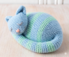 gatinUm gatinho amigurumi com listras cinzas e brancas, olhos bordados fechados como se estivesse dormindo, usado como peso de porta encostado em uma porta de madeira branca.ho-peso-de-porta-amigurumi-pronto